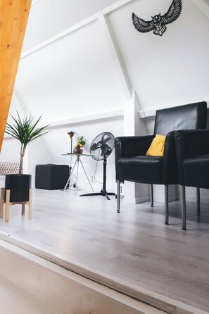 A modern living room with a black leather sofa, a small table with a lamp, a fan, and a potted plant. - Cosy studio in Hoorn perfect for a relaxing getaway (Hoorn)