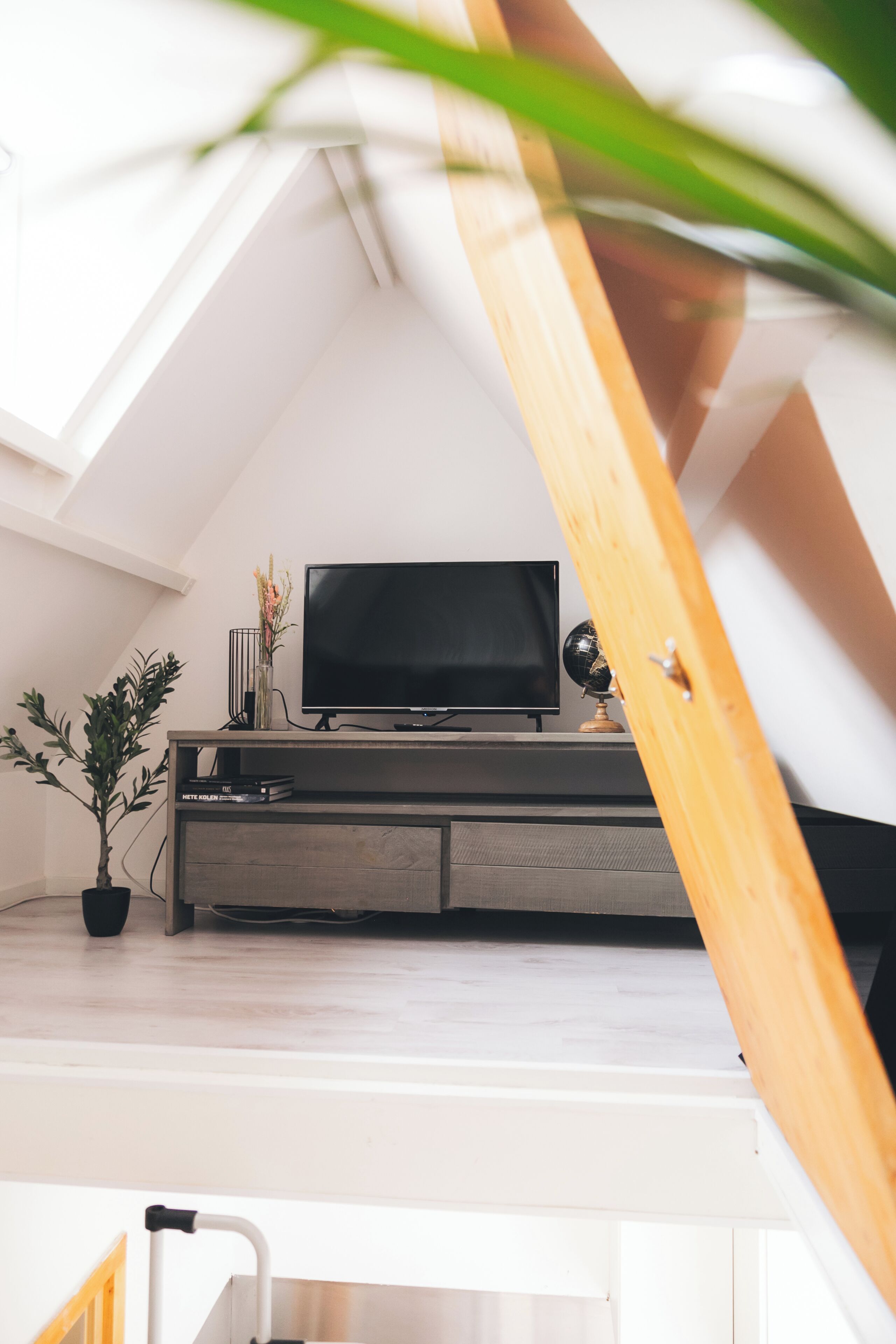 A modern loft bedroom with a sloped ceiling, a television on a wooden console, a globe, and a potted plant.