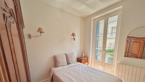 A bedroom with a bed, a nightstand, two wall-mounted lamps, a window with shutters, and a radiator. - Bright T2 Bordeaux | High ceilings & exposed stone | View of tree-lined square (Bordeaux)