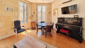A room with a piano, a dining table with chairs, a TV mounted on the wall, and large windows. - Bright T2 Bordeaux | High ceilings & exposed stone | View of tree-lined square (Bordeaux)