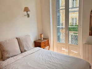 A bedroom with a bed, a nightstand, a lamp, and a view of a balcony with a railing. - Vrbo Property (Bordeaux)