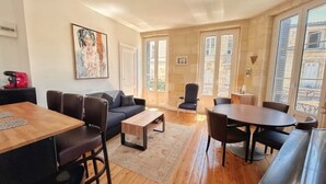 A modern living room with a sofa, coffee table, and dining table. There are two doors leading to a balcony. - Bright T2 Bordeaux | High ceilings & exposed stone | View of tree-lined square (Bordeaux)
