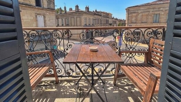 An outdoor balcony with a wooden table and chairs, a wrought iron railing, and a view of historic buildings. - Bright T2 Bordeaux | High ceilings & exposed stone | View of tree-lined square (Bordeaux)