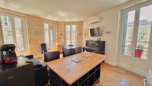 A spacious room with a wooden dining table, black chairs, a piano, and large windows. - Bright T2 Bordeaux | High ceilings & exposed stone | View of tree-lined square (Bordeaux)