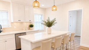 Uma cozinha moderna com armários brancos, uma ilha central com banquetas, iluminação pendente e um vaso de planta.