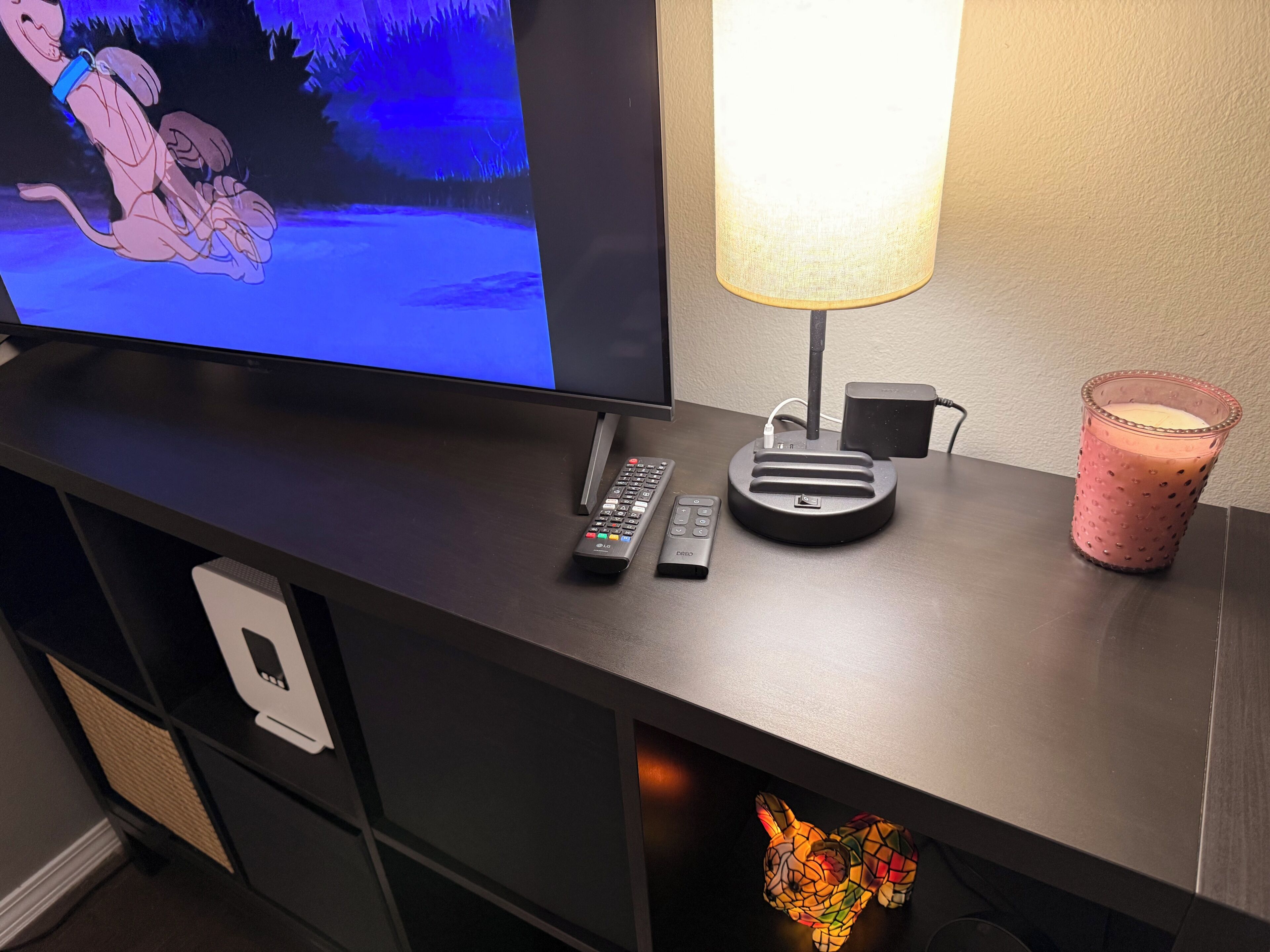 Una TV, telecomandi, una lampada, una candela e un oggetto decorativo su una scrivania.