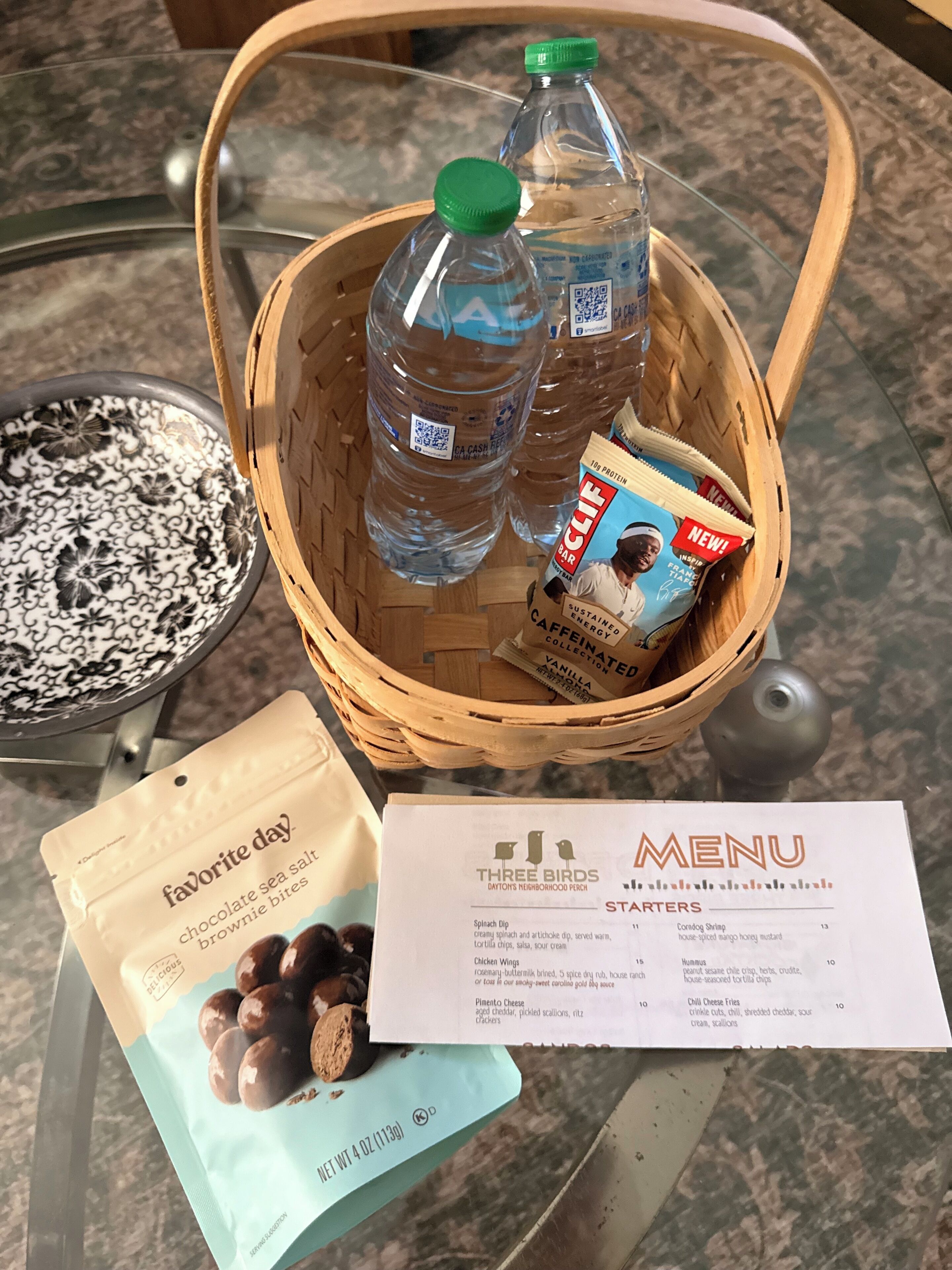 A basket with bottled water, a magazine, and a bag of snacks on a table.