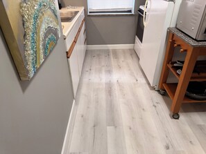 Een smalle keuken met een houten vloer, een witte koelkast, een magnetron en een houten plank met schoenen.