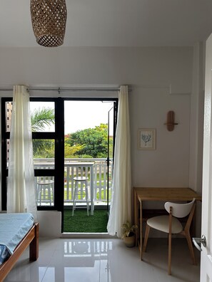 Ein Zimmer mit einem Bett, einem Holztisch, einem Stuhl, einer Pendelleuchte, Blick auf einen Balkon mit Geländer und einer Tür.