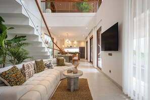 Een modern interieur met een trap, een zithoek met een bank en decoratieve kussens, een eethoek en een flatscreen televisie aan de muur.