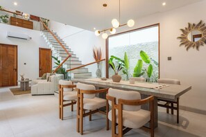 A modern living room with a dining area, a staircase, and a large window with a view. - Villa Albizia - 2BR private pool villa near Canggu (Mengwi)