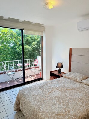 Quarto com cama, mesa de cabeceira, abajur, porta de vidro deslizante para área externa com mesa e cadeiras, e vista para vegetação.