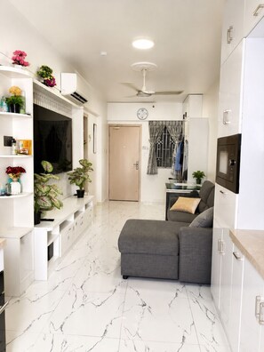 Una sala de estar moderna con un sofá gris, mueble de entretenimiento blanco y una zona de cocina con refrigerador y microondas.