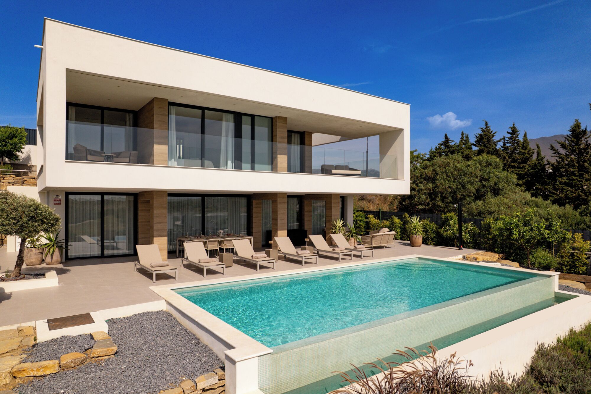 A modern two-story house with a swimming pool, outdoor seating, and a view of trees and mountains.