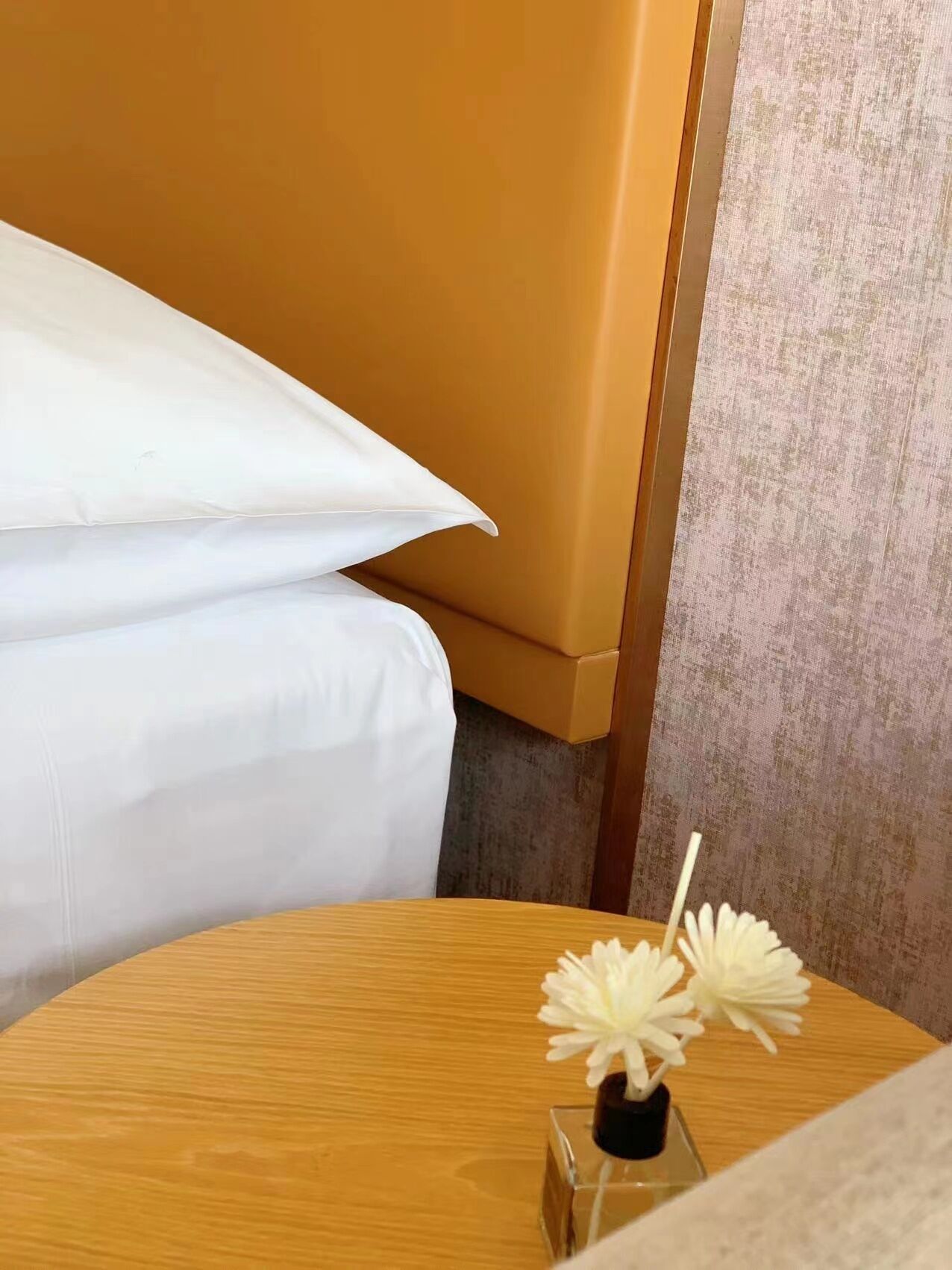 A hotel room with a bed, a wooden side table with a vase of white flowers, and a textured wall.