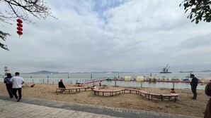 Una playa con un paseo marítimo pavimentado, una zona arenosa con bancos y vistas al mar y barcos a la distancia.