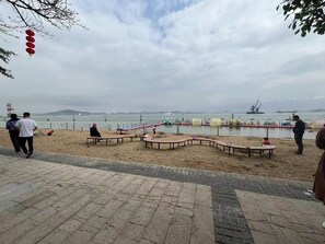 A beach with a paved walkway, a sandy area with benches, and a view of the sea and distant ships. - Vienna International Hotel (Guangzhou)