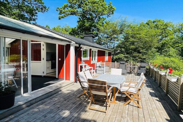A wooden deck with outdoor seating, a glass door entrance, and a red building in the background. - 4 star holiday home in KARLSHAMN (KARLSHAMN)