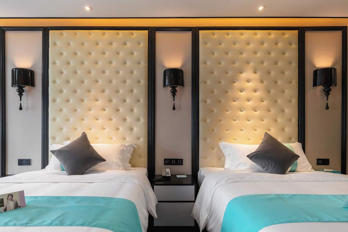 Habitación de hotel con dos camas, cada una con cabecero capitonado, ropa de cama blanca y almohadones color turquesa. Hay mesitas de noche con lámparas negras y una mesita auxiliar pequeña entre las camas.