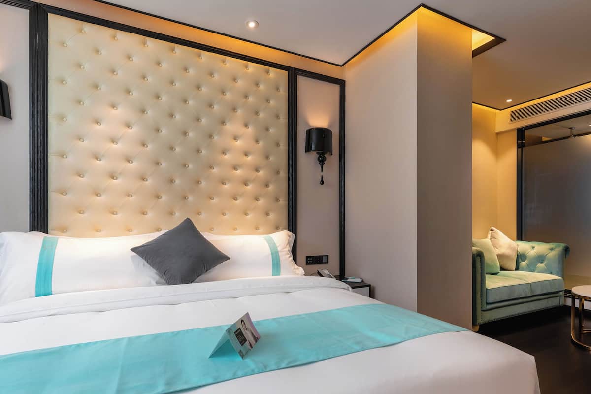 Habitación de hotel con una cama grande, un cabecero capitonado, una mesita de noche, una lámpara y un sillón tapizado en verde.