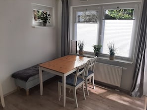 Una habitación con una mesa de madera, sillas blancas, un banco y plantas en macetas junto a las ventanas.