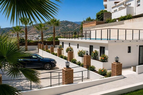 Un edificio moderno con un automóvil estacionado frente a él, rodeado de palmeras y vistas a un paisaje montañoso.