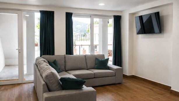 A living room with a beige sectional sofa, a flat-screen TV mounted on the wall, and a sliding glass door leading to an outdoor area.