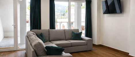 Una sala de estar con un sofá seccional color beige, un televisor de pantalla plana montado en la pared y un ventanal corredero que da a un área exterior.