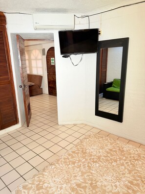 Ein Zimmer mit Fliesenboden, einem Bett mit gemustertem Überwurf, einem an der Wand montierten Fernseher und einem Spiegel, der einen grünen Sessel reflektiert.