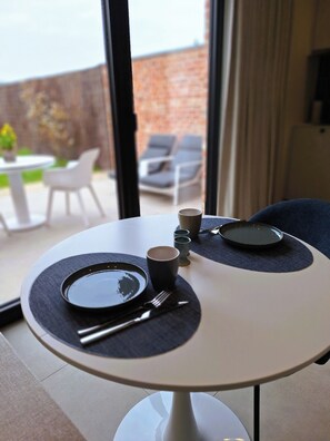 Una mesa de comedor con un plato, una taza y cubiertos, una silla, y vistas a una pared de ladrillo y sillones de exterior.