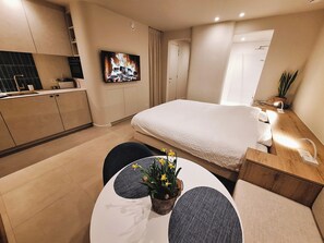 Een moderne hotelkamer met een bed, een klein eettafeltje met een plant in pot, een televisie en een kitchenette.