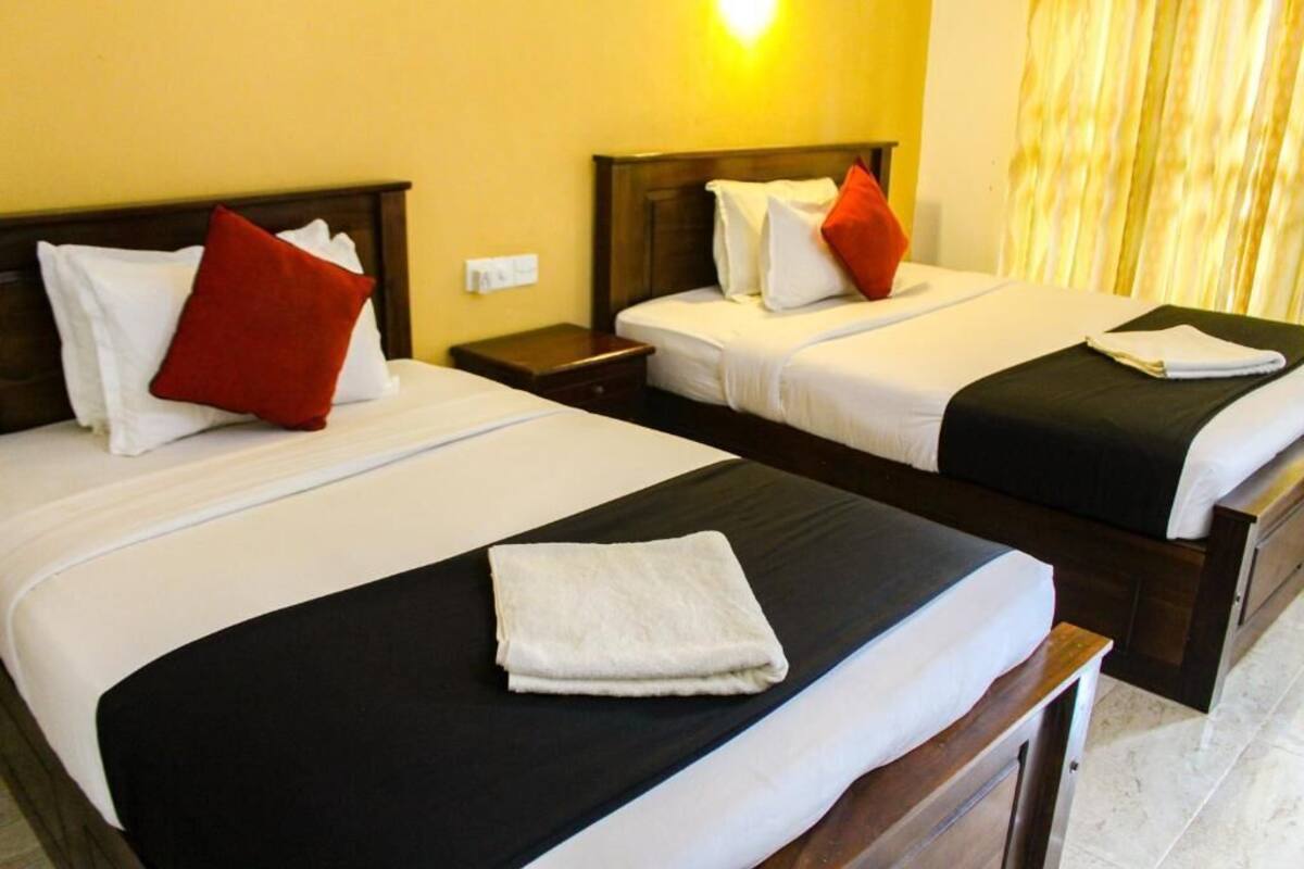 Two beds with white linens and red pillows, a wooden headboard, a nightstand, and a window with curtains.