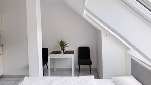 Une petite chambre moderne, équipée d’un lit, d’une table avec une plante, de deux chaises et d’une fenêtre.