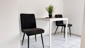 Une petite pièce moderne avec un sol carrelé de couleur blanche, deux chaises noires, une table blanche et une plante en pot.