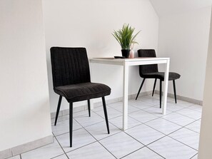 Una habitación pequeña y moderna con piso de baldosas blancas, dos sillas negras, una mesa blanca y una planta en maceta.