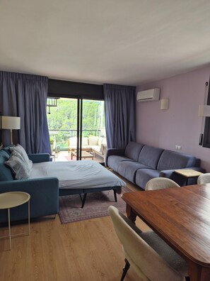 A modern living room with a sofa, a bed, a dining table, and a view of greenery outside.