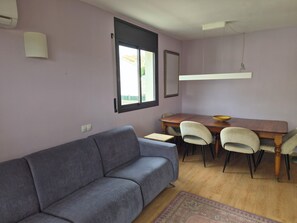 A living room with a grey sofa, a wooden dining table with chairs, a patterned rug, and a window with blinds.