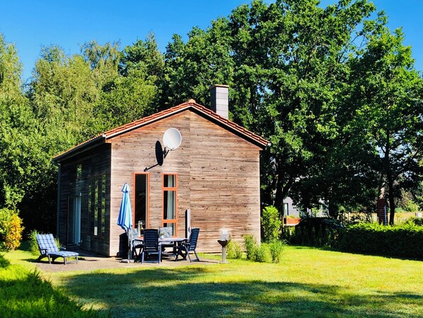 A wooden cabin with a satellite dish, a covered patio with a table and chairs, and a grassy yard. - Vrbo Property (Fuhlendorf)