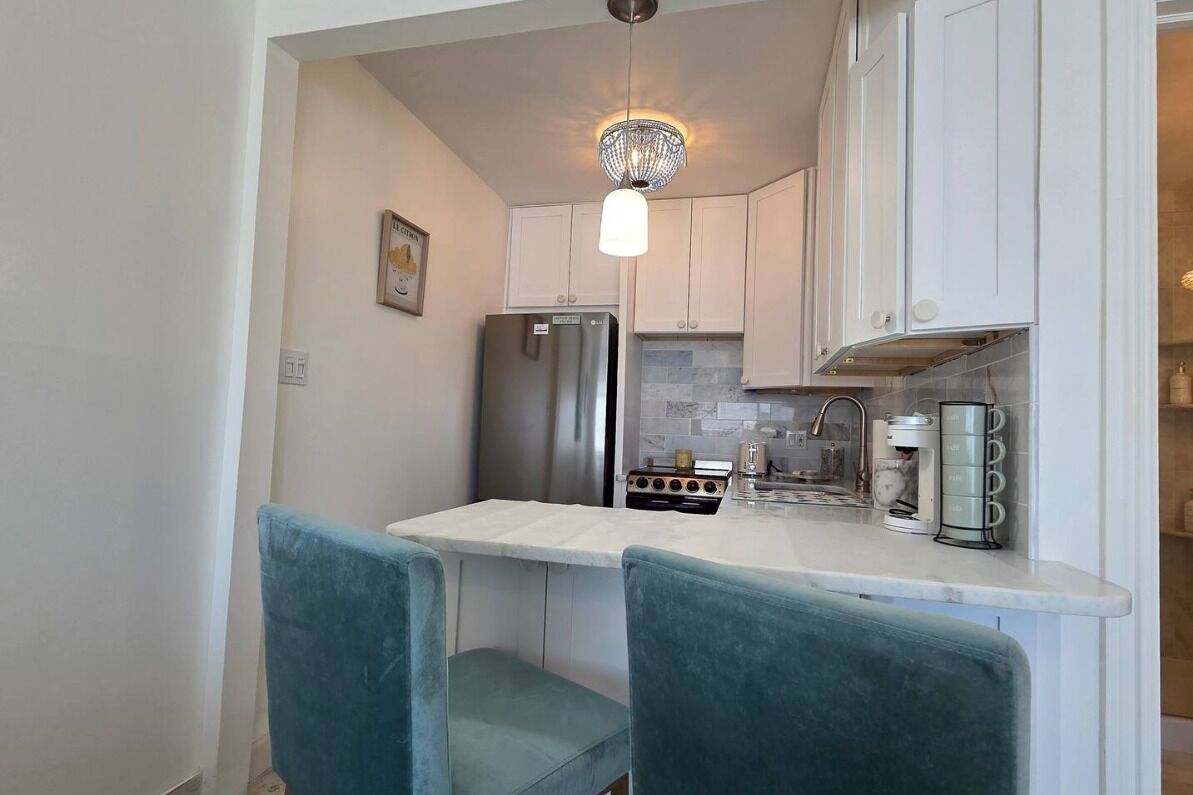 A compact kitchen with a refrigerator, stove, and coffee maker. There are two chairs and a pendant light.