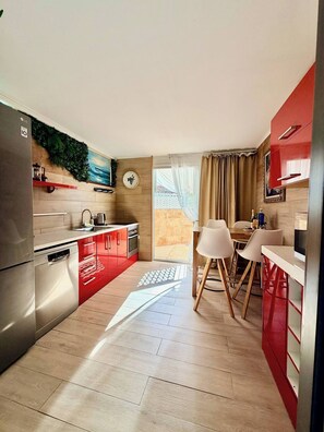 Een moderne keuken met een koelkast, fornuis en vaatwasser, een eethoek met een tafel en stoelen, en een uitzicht door een raam met gordijnen.