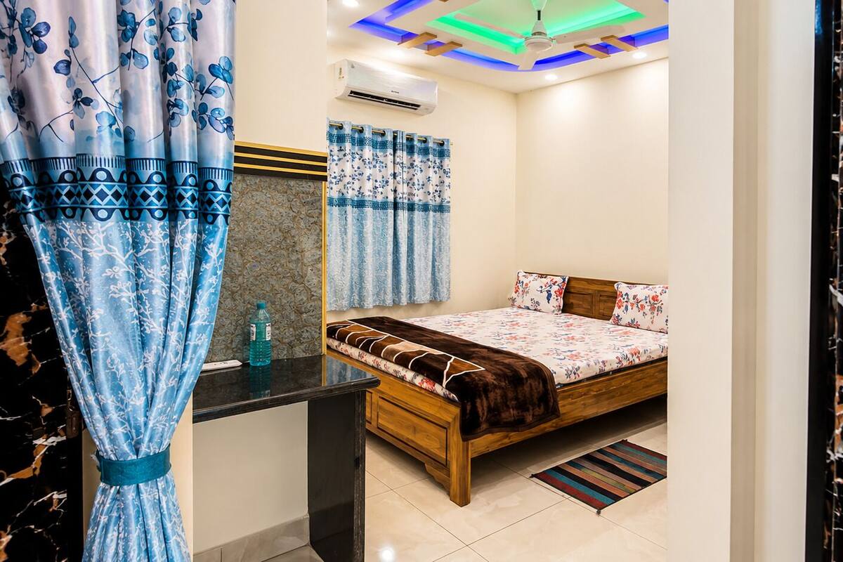 A bedroom with a bed, curtains, an air conditioner, a ceiling light, and a wooden nightstand.