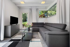 A modern living room with a sofa, a glass coffee table, a flat-screen TV, and a view of greenery through a window.