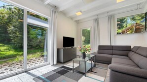 Ein modernes Wohnzimmer mit einer Couch, einem Flachbildfernseher, einem Glastisch und großen Fenstern mit Blick auf Grünflächen.