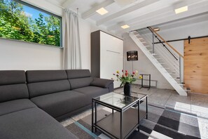 A modern living room with a sectional sofa, a glass coffee table, a staircase, and a view of greenery outside.