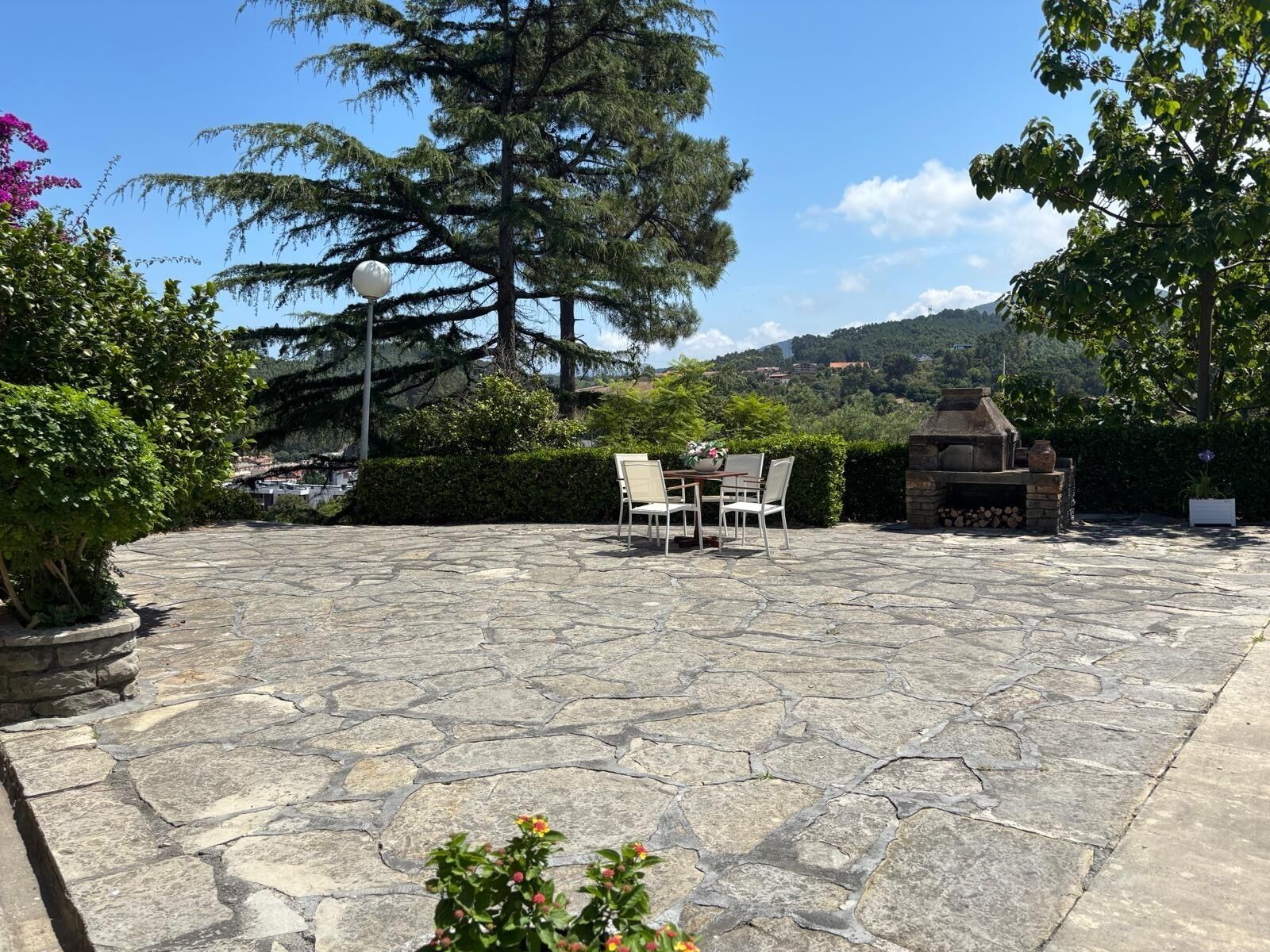 Un patio de piedra con mobiliario exterior, una parrilla y un gran árbol.