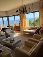 Uma sala de estar ampla com uma grande janela com vista para o mar, um candelabro e um conjunto de sofás.