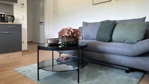 A modern living room with a grey sofa, a glass coffee table with a flower vase, and a kitchen area in the background. - FarawayHomes Studio Leoben #P72 (Donawitz)