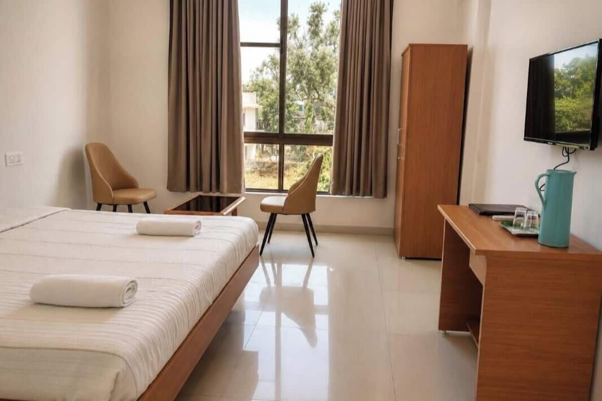 A hotel room with a bed, two chairs, a desk, a television, and a window with curtains.