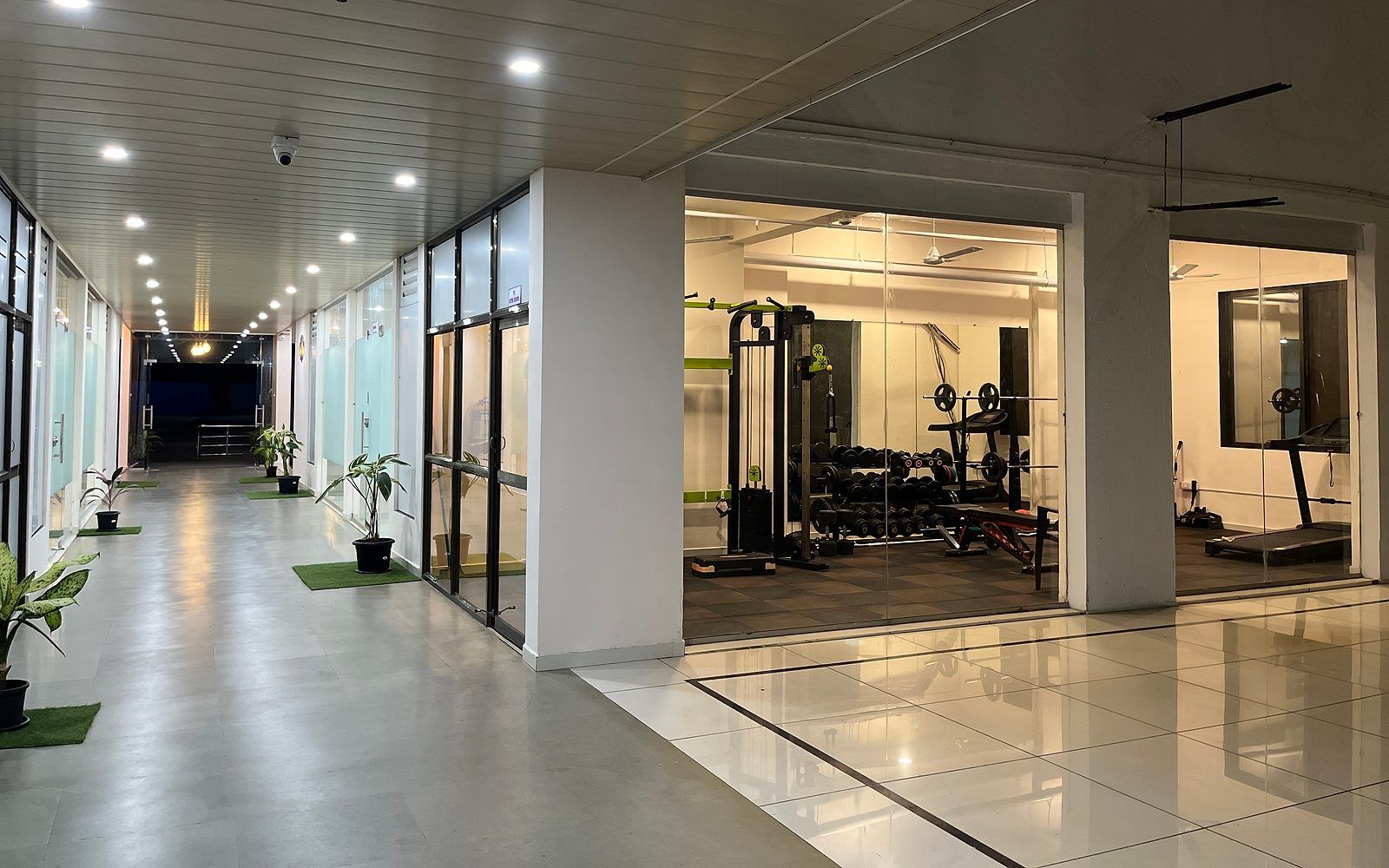 Un gimnasio con equipamiento de ejercicio, un pasillo con plantas en macetas e iluminación empotrada en el techo.