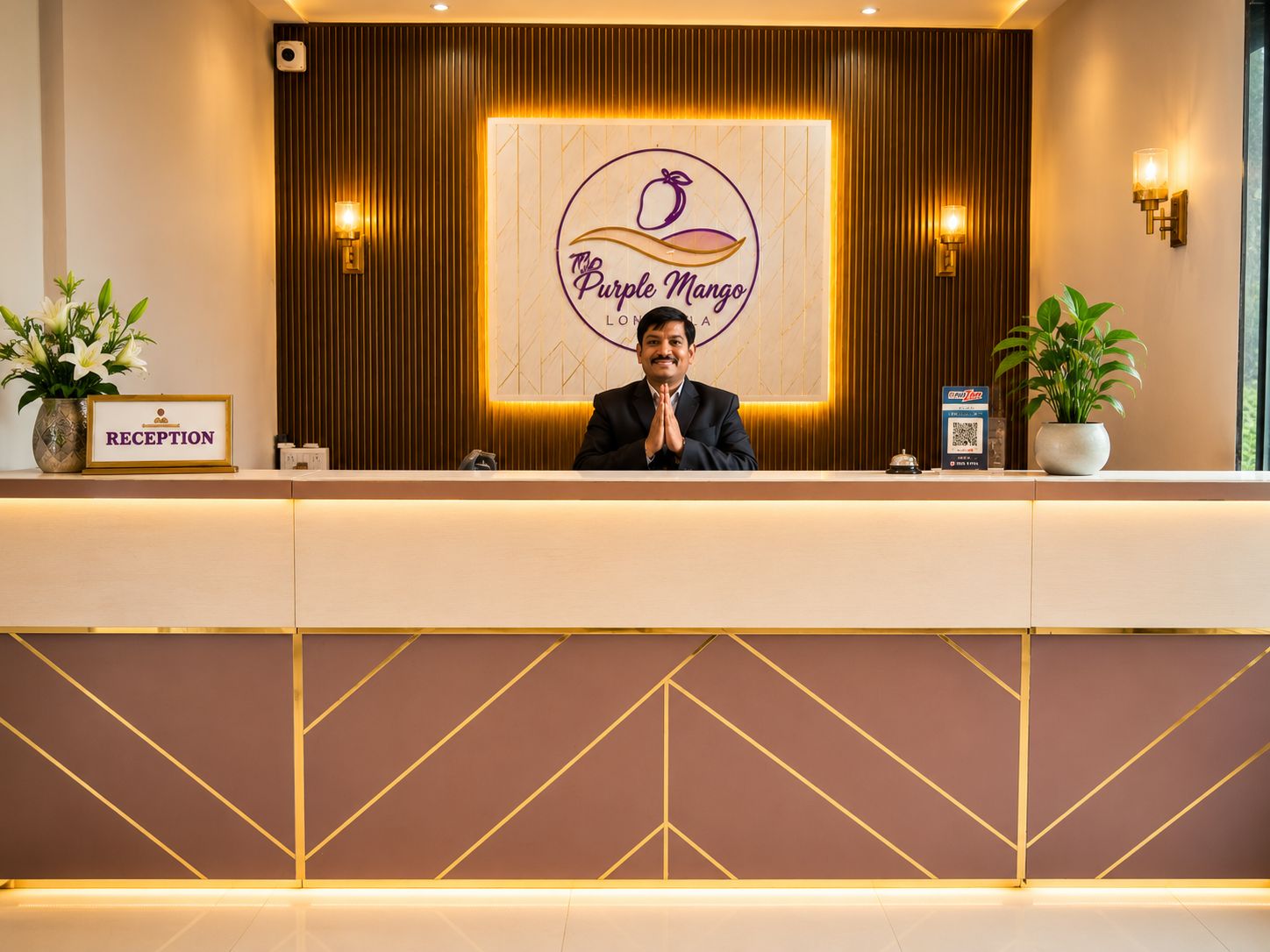 A reception desk with a sign reading 'The Purple Mango' and a 'RECEPTION' sign. There are two potted plants on either side of the desk.